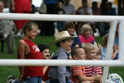 Group of children smiling behind a metal railing; one child wears a cowboy hat in the center.